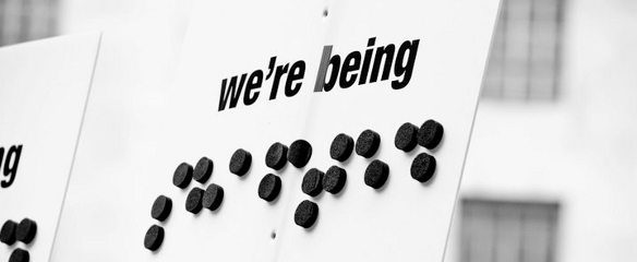 Black and white close up photo of a 2011 banner, "We're being Shafted" by the government (shafted as braille letters)